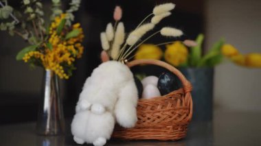Yakın plan. Paskalya kutlaması. Dairedeki masada içinde bahar çiçekleri olan cam bir vazo, bir Paskalya tavşanı ve içinde Paskalya yumurtaları olan bir sepet var. Bir kadın el yapımı bir Paskalya kartı koyar.