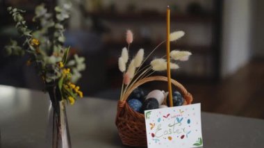 Yakın plan. Mutlu Paskalyalar. Yanan bir mum, içinde bahar çiçekleri olan cam bir vazonun yanında paskalya yumurtaları bulunan süslü bir sepetin içinde duruyor. Sepetin yanında duran el yapımı Paskalya kartı.