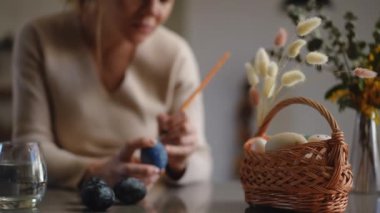 Ağır çekim. Yakın plan. Bir kadın oturma odasındaki bir masaya oturur ve Paskalya yumurtasındaki suluboyayı boya fırçasıyla süsler. Paskalya kutlaması için hazırlıklar