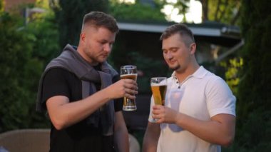 Ağır çekim. İki genç sakallı adam ellerinde bira bardaklarıyla dışarıda duruyor. Erkekler bira bardaklarını tokuşturur ve gülümseyen kameraya bakarlar.