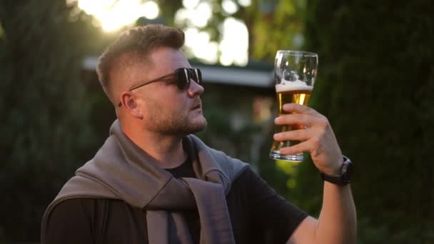 スローモーション クローズアップ サングラスのスタイリッシュな若いひげの男はビールを味わい この飲み物の肯定的な評価を与えます 男は夏の夕方に屋外でビールグラスを持って立っている — ストック動画