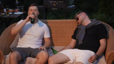 Açık havada bir yaz akşamında hasır bir kanepede oturan iki adam eğleniyor. Bir adam bira içiyor ve elinde bir şişe birayla uyuyakalan güneş gözlüklü arkadaşına gülümsüyor.
