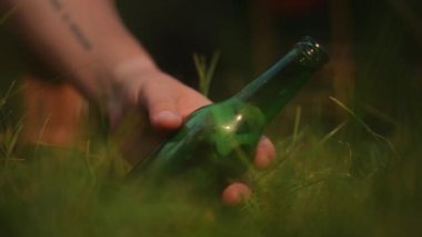 Yakın plan. Bitmemiş bir bira şişesi, dövmeli genç bir adamın elinden çimenlere düşüyor.