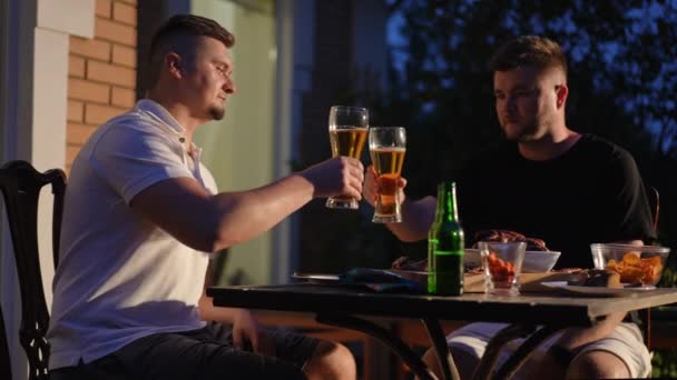 2人の男性は 夏の夕方にグリルソーセージとチップで木製のテーブルでビールのメガネで屋外に座っています 男性はメガネを飲んでビールを飲む — ストック動画