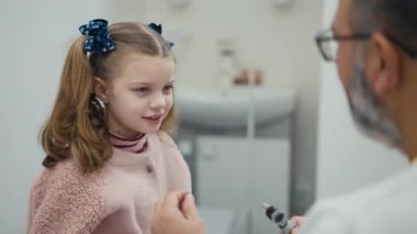 Bir pediatrist, modern bir klinikte muayene koltuğunda oturan küçük bir kızın kulağını görsel olarak muayene eder. Erkek bir doktor bir çocuğun kulağını muayene eder. Kız gülümsüyor ve onu okşuyor.