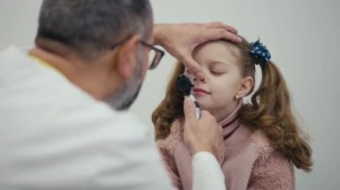 Yakın plan. Bir erkek çocuk doktoru, modern bir klinikte muayene koltuğunda oturan küçük bir kadın hastanın burun boşluğunu görsel olarak muayene eder.