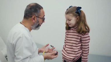 Erkek bir çocuk doktoru, modern bir klinikte muayene koltuğunun yanında duran küçük bir kız hastayı muayene ederken steteskop kullanır. Doktor çocukla konuşur ve nefes alışını dinler.