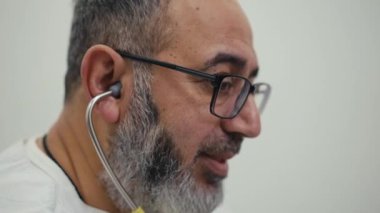 Gözlüklü sakallı bir adamın yüzünü yakından çek. Kulağında steteskopla bir hastayla konuşuyor.