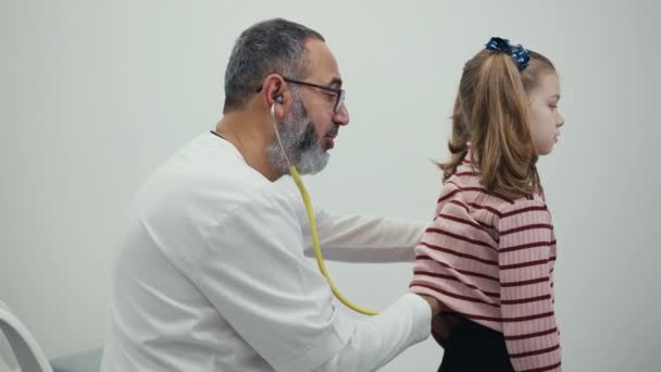 一位男医生把听诊器放在一位小女病人的后背 在一家现代诊所的预约中听孩子们的呼吸 — 图库视频影像