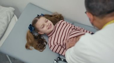 Omzunun üstünden. Küçük bir kız hasta sırt üstü tıbbi muayene koltuğunda yatıyor ve karnını bir çocuk doktoru muayene ederken gülüyor. Erkek bir doktor karnını muayene ediyor.