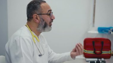 Boynunda steteskop olan bir doktorun profili. Ofisindeki bir masada oturmuş, hastasıyla konuşurken elleriyle el kol hareketi yapıyor.