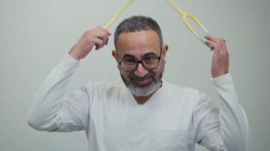 Sakallı ve gözlüklü bir erkek doktor kameraya bakıyor, gülümsüyor ve boynuna steteskop takıyor.