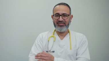 Gözlüklü, boynunda steteskop olan, beyaz arka planda duran sakallı bir erkek doktorun portresi. Adam kameraya bakarken gülümsüyor ve kollarını göğsünde kavuşturuyor.