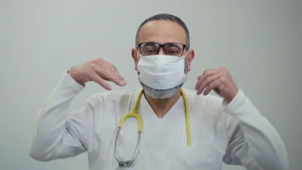 首に刺繍をした男性医師は 屋内の白い背景に立って 顔に医療マスクを置きます 男は彼の手で顔のマスクを調整し 見える — ストック動画