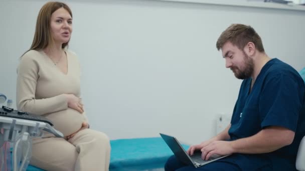 医師による定期検査中の妊婦 妊娠中の女性は診察ソファに座り 笑顔で椅子に座ってすべてを書く男性医師とコミュニケーションをとります — ストック動画