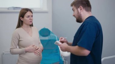 Bir doktor, jinekoloji kürsüsünün yanında duran hamile bir kadına tavsiyelerde bulunur. Kadın ellerini karnının üstüne koyup doktorla aynı fikirde başını sallıyor.