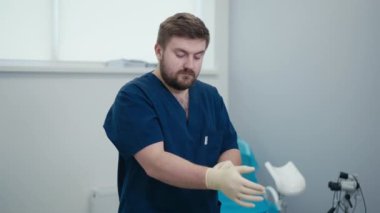 Mavi tıbbi üniformalı sakallı bir doktor ellerine tıbbi eldiven takıyor ve ofisinde jinekoloji sandalyesinin yanında duran kameraya bakıyor.