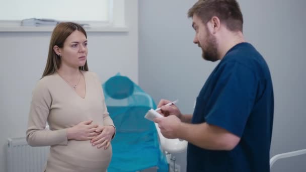 医師は婦人科の椅子の近くに立っている間に妊婦に勧告を与えます 女性は彼女の腹の上に彼女の手を握り 医者と同意して彼女の頭をノック — ストック動画