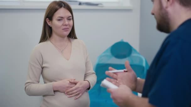 妊娠中の女性は 彼の勧告の間に医師のオフィスに立っている間 微笑んでいます 女性は彼女の胃に手を握り 医者と同意して彼女の頭をノックします — ストック動画