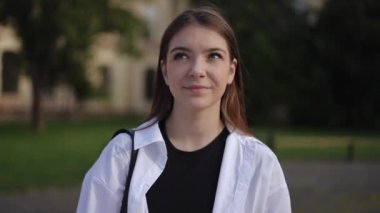 Yakın plan. Güzel bir kız, bir yaz günü, yeşil bir parkın ve eski bir binanın zemininde duran kameraya ve tatlı gülümsemelere bakar.