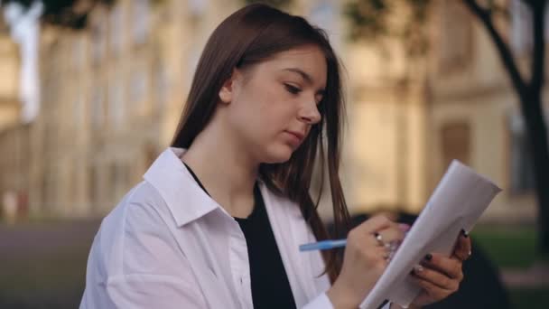夏天的一天 一个女孩坐在研究所的公园里 手里拿着笔在笔记本里写字 — 图库视频影像