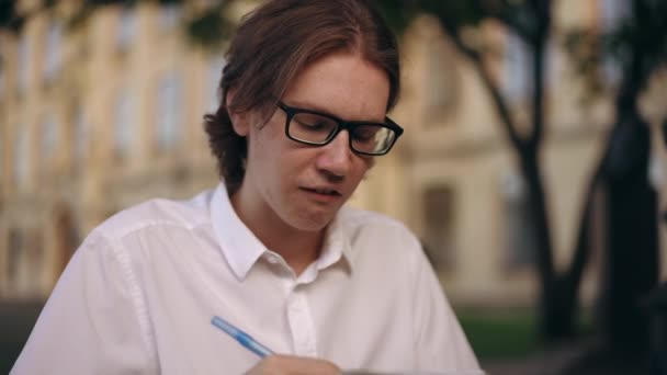 一个戴眼镜的年轻人拿着笔在笔记本里写字 夏天的一天 他站在室外一座大楼的背景下 — 图库视频影像