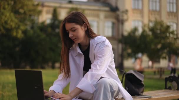 慢动作一个夏天的晚上 在研究所的公园里 一个女孩坐在外面的长椅上 在笔记本电脑前工作 这个女孩看着练习本 在笔记本电脑上打字 — 图库视频影像