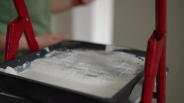 Ağır çekim. Yakın plan. Yeşil tulumlu tanınmayan bir kadın, katlanan bir merdivenin basamağında duran beyaz boyalı geniş bir boya silindiri yuvarlıyor..