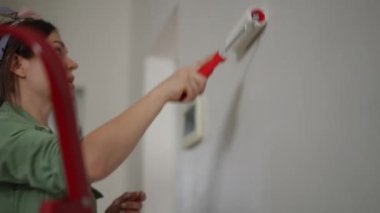Yeşil önlüklü bir kadın ustaca resim yapar, beyaz boyalı geniş bir boya rulosunu yuvarlar, koridor duvarı, kırmızı katlanır merdiven zemininde durur..