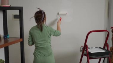 Dikiz aynası. Yeşil tulumlu bir kadın, beyaz boyalı geniş bir boya silindirini yuvarlayarak bir duvar boyuyor. Bir kadın kırmızı bir merdivenin yanında duruyor.