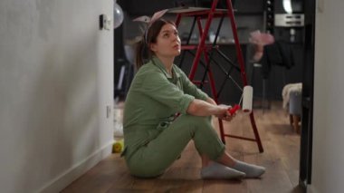 Ağır çekim. Yeşil önlüklü yorgun bir kadın kırmızı bir merdiven yakınındaki ahşap bir zeminde oturuyor ve alnından terini eliyle siliyor. Bir kadın elinde beyaz boyayla bir boya silindiri tutuyor.
