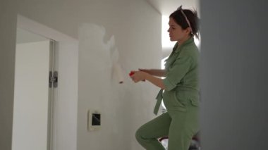 Yeşil tulumlu hamile bir kadın beyaz boyalı bir boya rulosuyla bir duvarı boyuyor, bir apartmanın koridorundaki merdiven üzerinde duruyor.