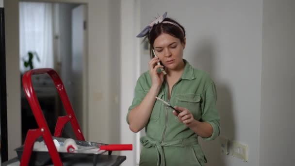 一位穿着绿色工作服的漂亮女人一边站在公寓走廊一边刷墙 一边用手机接听电话 一个女人在用手机聊天 面带微笑 — 图库视频影像