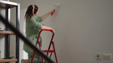 Ağır çekim. Yeşil tulumlu hamile bir kadın merdiven üzerinde duran bir boya silindiriyle koridor duvarını boyuyor. Kadın yorgun ve alnından terini eliyle siliyor. DIY duvarı