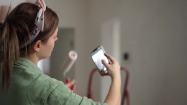 Elinde boya silindiri olan yeşil tulumlu bir kadın cep telefonu kamerasıyla fotoğraf çekiyor. Dairesindeki duvarları nasıl boyuyor?