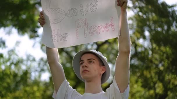若い男は 彼の頭の上に惑星を保存する碑文付きのポスターを保持し 夏の日に市立公園で屋外に立っています — ストック動画