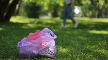 Ön planda, bir yaz günü şehir parkında yeşil çimlerin üzerinde plastik bir çöp torbası bulunur. Tanınmayan bir adam çimlere saçılmış çöpleri topluyor.