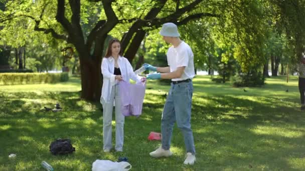 屋外の都市公園でゴミを掃除する 少女はゴミを持ったビニール袋を手に持っている 若い男 太陽の帽子とゴムの手袋で 草の上に散らばったゴミを集めて置く — ストック動画