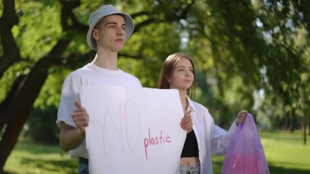 抗议对塑料说不 夏天的一天 一个男人和一个女孩在公园里一动不动地站在外面 这个家伙手里拿着一张没有塑料海报 而那个女孩手里拿着一个塑料袋 — 图库视频影像