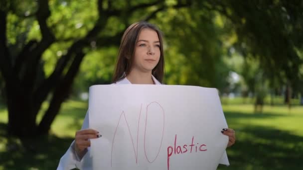 女の子は 市立公園の屋外で夏の日に立って 彼女の手にプラスチックのポスターを持っていません 若い女性が微笑んでいる プロテスタントはプラスチックにノーと言う — ストック動画
