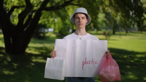 太陽の帽子をかぶった若者はカメラを見て 市の公園で晴れた夏の日に立っている間 手元にプラスチックのポスターを置いていません 環境保護キャンペーン — ストック動画