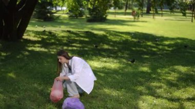 Bir kız güneşli bir yaz gününde şehir parkında yürürken çimenlere saçılmış çöpleri plastik bir torbaya koyar.