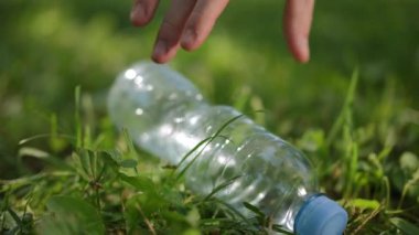 Ağır çekim. Yakın plan. Bir adamın eli, şehir parkındaki çimlerin üzerinde duran boş bir plastik şişeyi kaldırır.