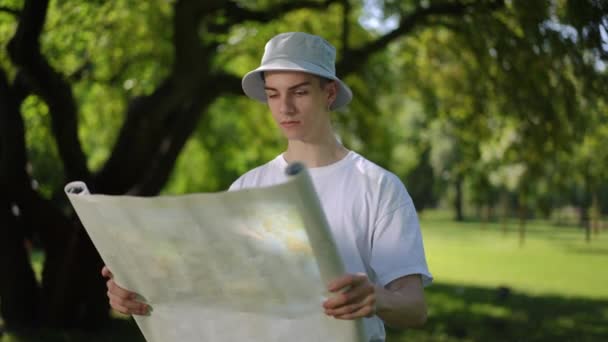 スローモーション 太陽の帽子をかぶった男は 手元に展開された地図を持ち 屋外の公園で晴れた夏の日を過ごしながら周りを見回します — ストック動画