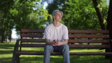 Güneş şapkalı bir adam gergin ve telaşlı bir şekilde elinde cep telefonu ekranına bakarken güneşli bir yaz gününde şehir parkında bir bankta oturuyor.