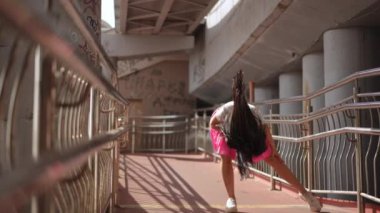 Ağır çekim. Güneş gözlüklü uzun saçlı güzel bir kız şehrin yaya köprüsünün altında zarif bir sokak dansı yapıyor. Kız dans ediyor, kameraya bakıyor ve kollarını kaldırıyor.