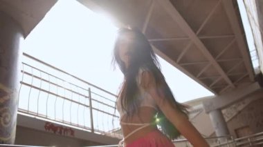 Ağır çekim. Düşük açılı atış. Uzun saçlı bir kız kameraya bakar ve kollarını zarif bir şekilde hareket ettirir, sokak dansı yaparken, şehrin yaya köprüsünün altında güneşin önünde dururken.