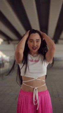 Dikey video. Siyah uzun saçlı sevimli bir kız kameraya poz veriyor. Kız kameraya bakıyor, saçını düzleştiriyor ve zarif bir şekilde ellerini hareket ettiriyor. Şehrin altında bir yaz günü ayakta duruyor.