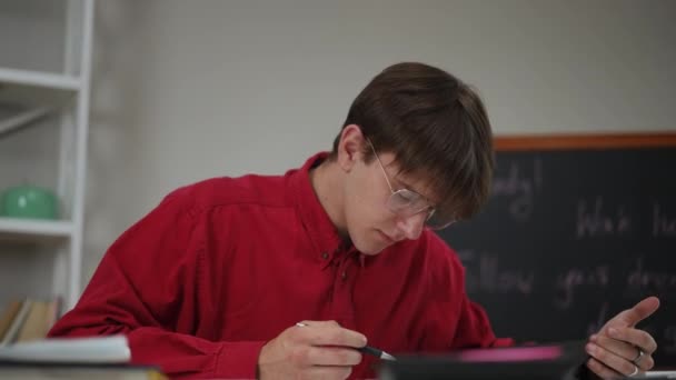 メガネの男性学生と赤いシャツ 部屋のテーブルに座って ノートパッドを開き データを見て 計算機のデータを数え 結果をペンでエクササイズブックに書き込む — ストック動画