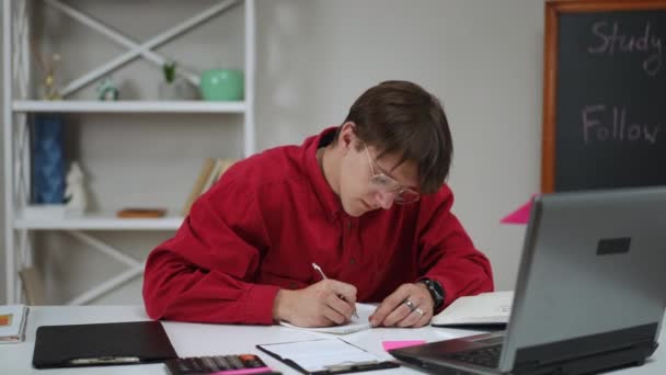 一个戴眼镜的年轻人看着笔记本电脑屏幕 用笔在笔记本上写字 用桌上的课堂笔记核对数据 一个学生坐在桌旁准备考试 — 图库视频影像